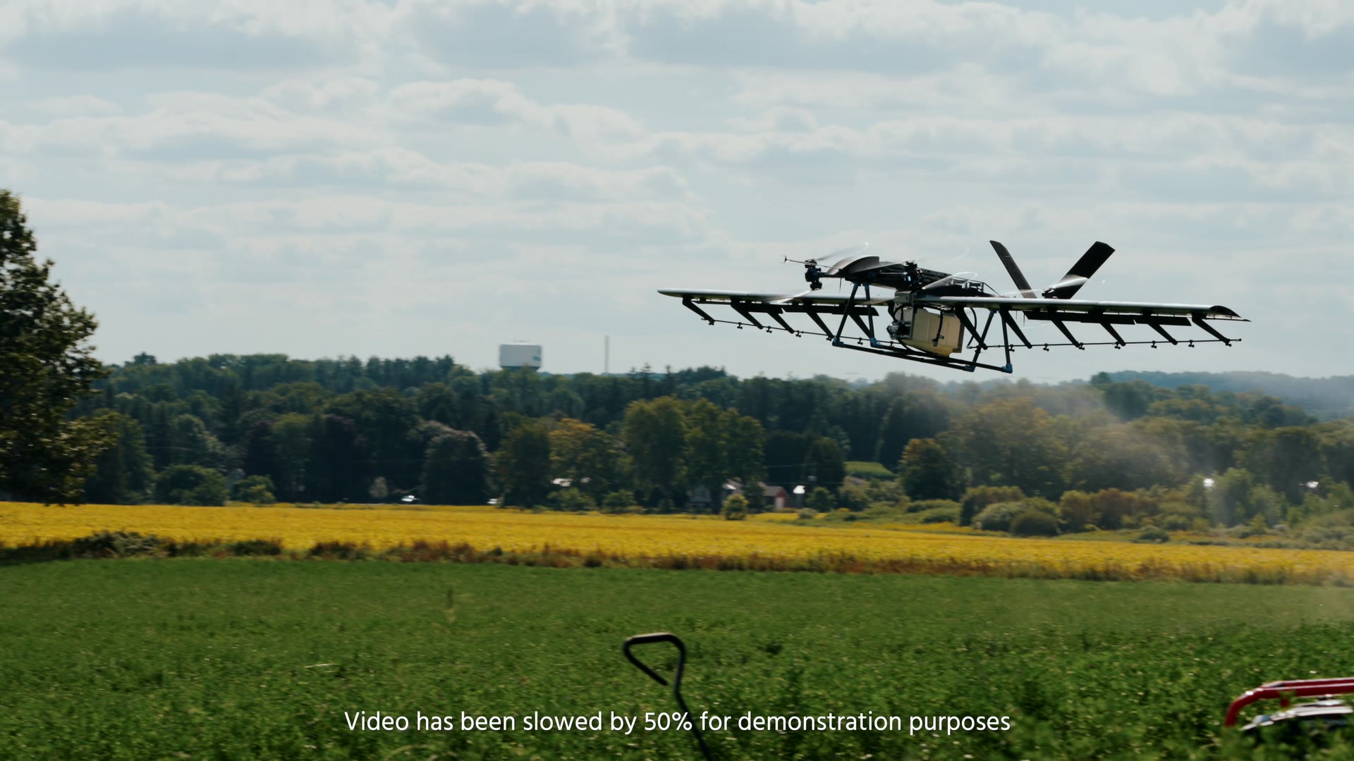 The U9 Spray in flight over a field, laying down a spray pass, with a treeline and a strip of flowering crop behind it.